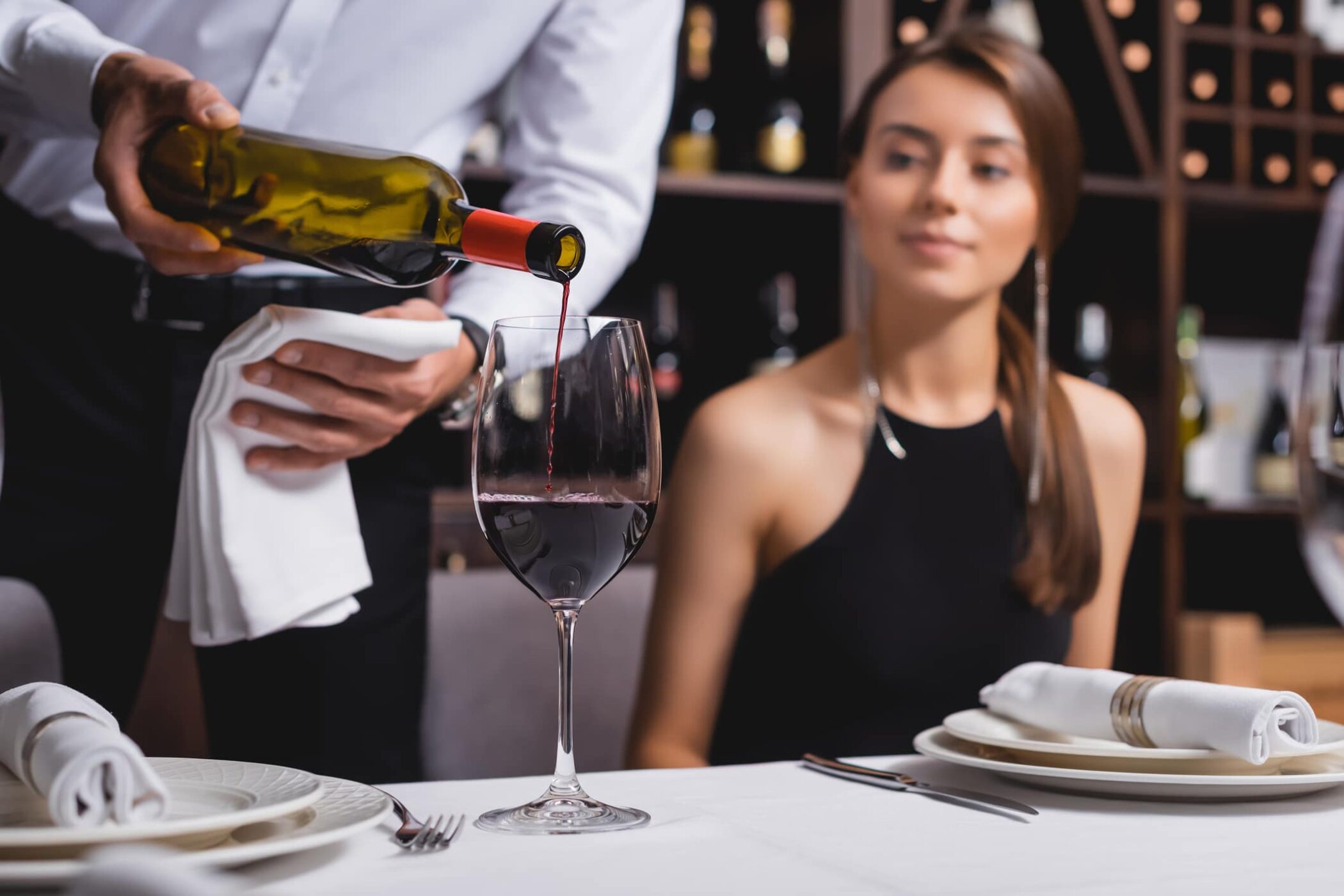 Selective focus of a wine steward pouring wine near woman at table in restaurant