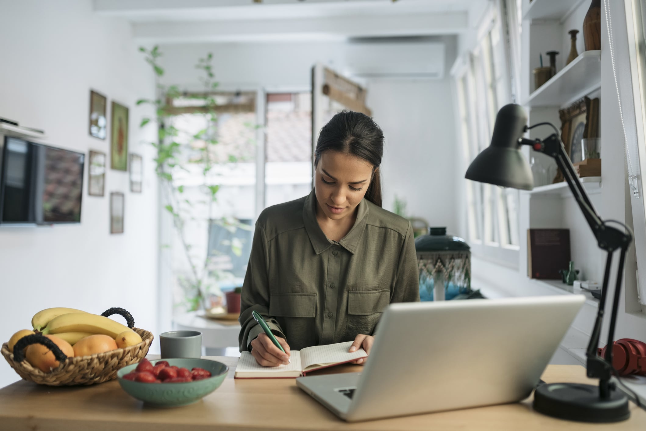 Unemployed young woman looking for a job online from home using laptop and writing down the notes in her notebook